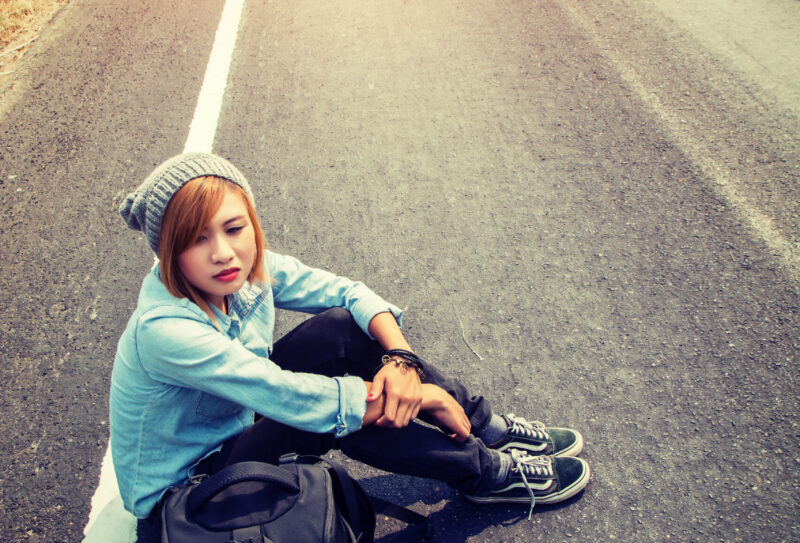 Rear view of a young woman hitchhiking carrying backpack sitting on the road